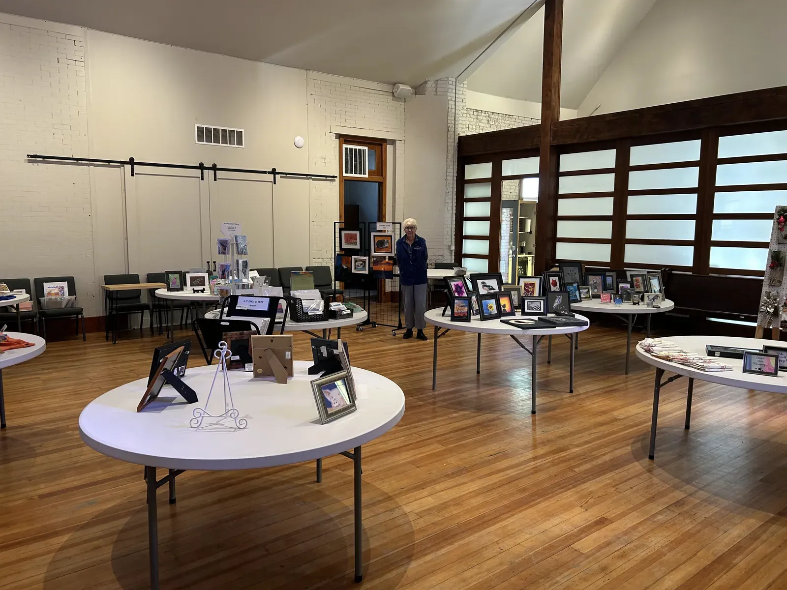 Framed artwork displayed on round tables across the hall's wood floor during a community art show