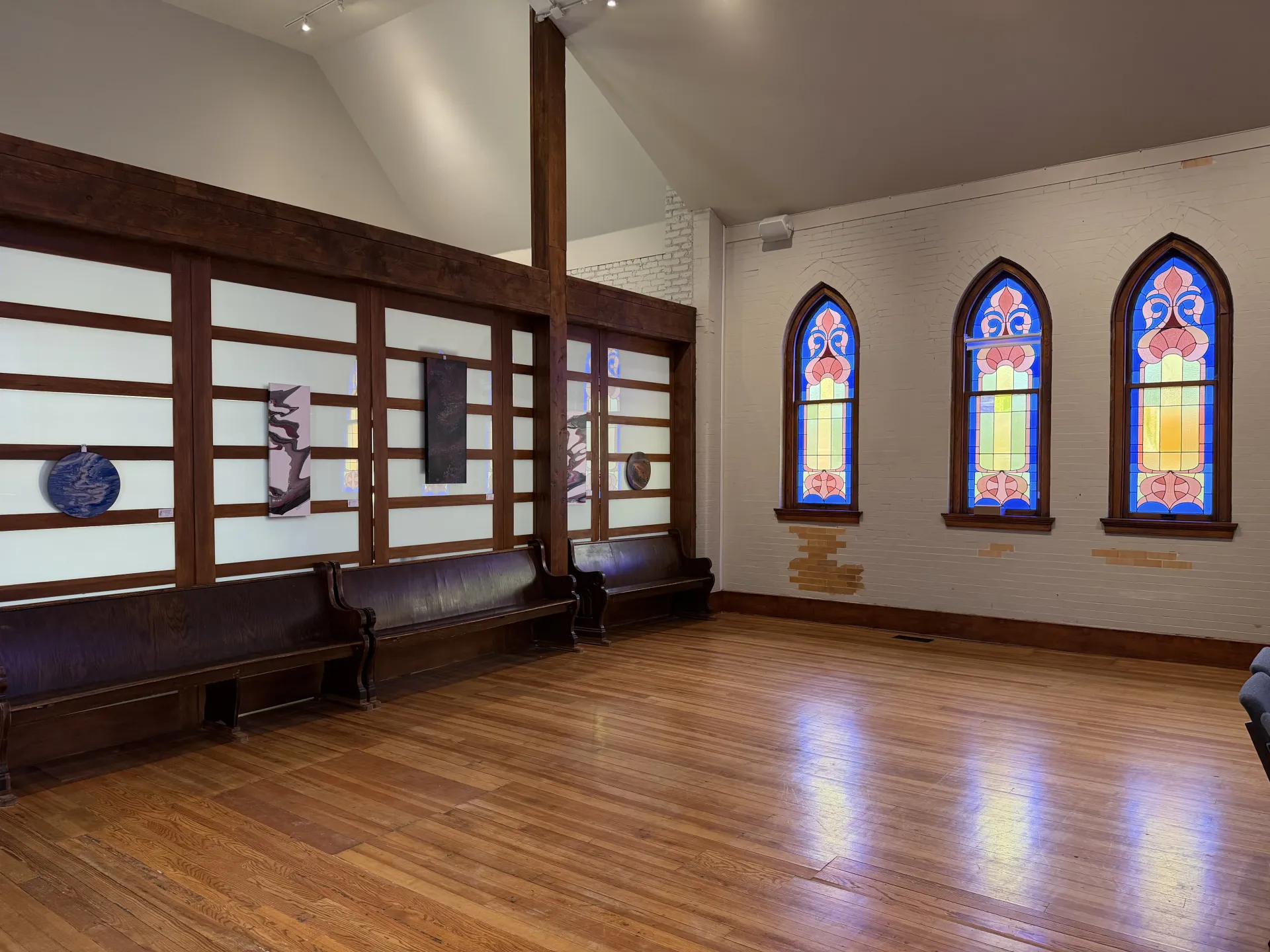 Sunlit hall with artwork displayed along the partition wall and three arched stained-glass windows lighting the wood floor