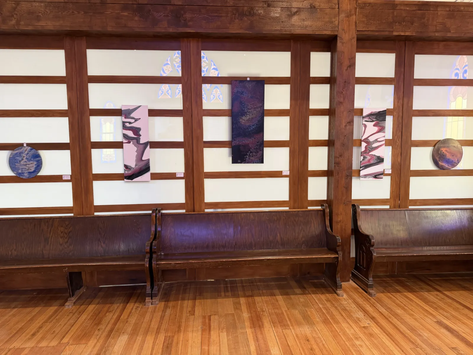 Framed abstract works on the partition wall above a row of antique wooden pews, stained glass glowing through the panels