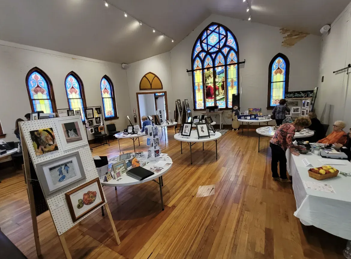Visitors browse framed artwork on easels and round tables during an art show beneath the hall's arched stained-glass windows