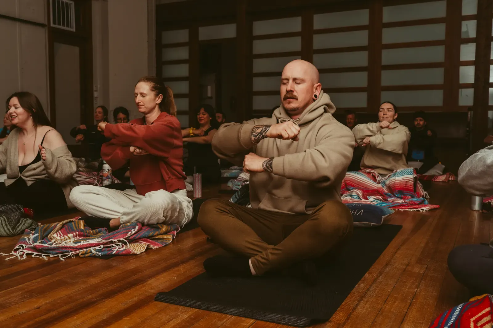 A breathwork class practices seated arm movements together on the hall's wood floor