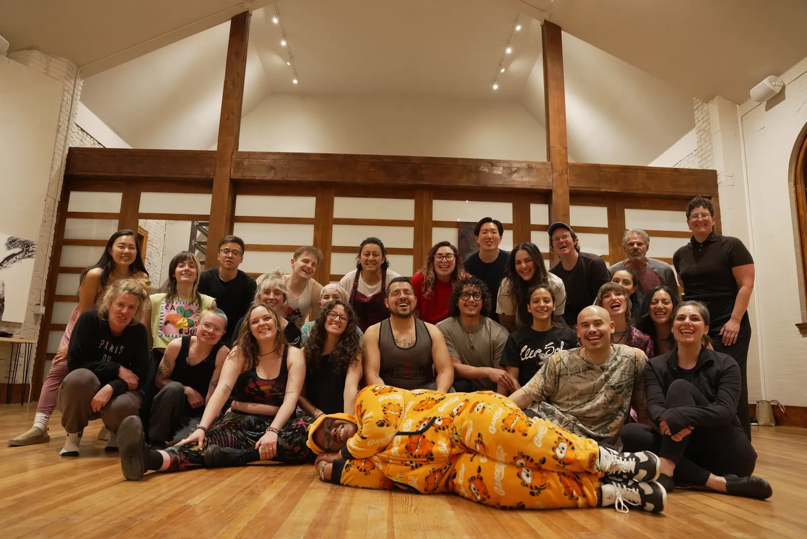 Class members sit in a circle on the wood floor for a discussion, reflected in the studio's rollaway mirror