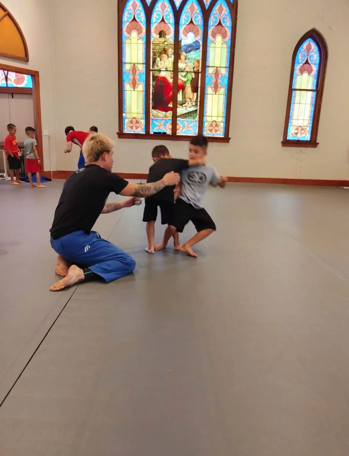An instructor coaches two young students practicing grappling on the full-coverage floor mat