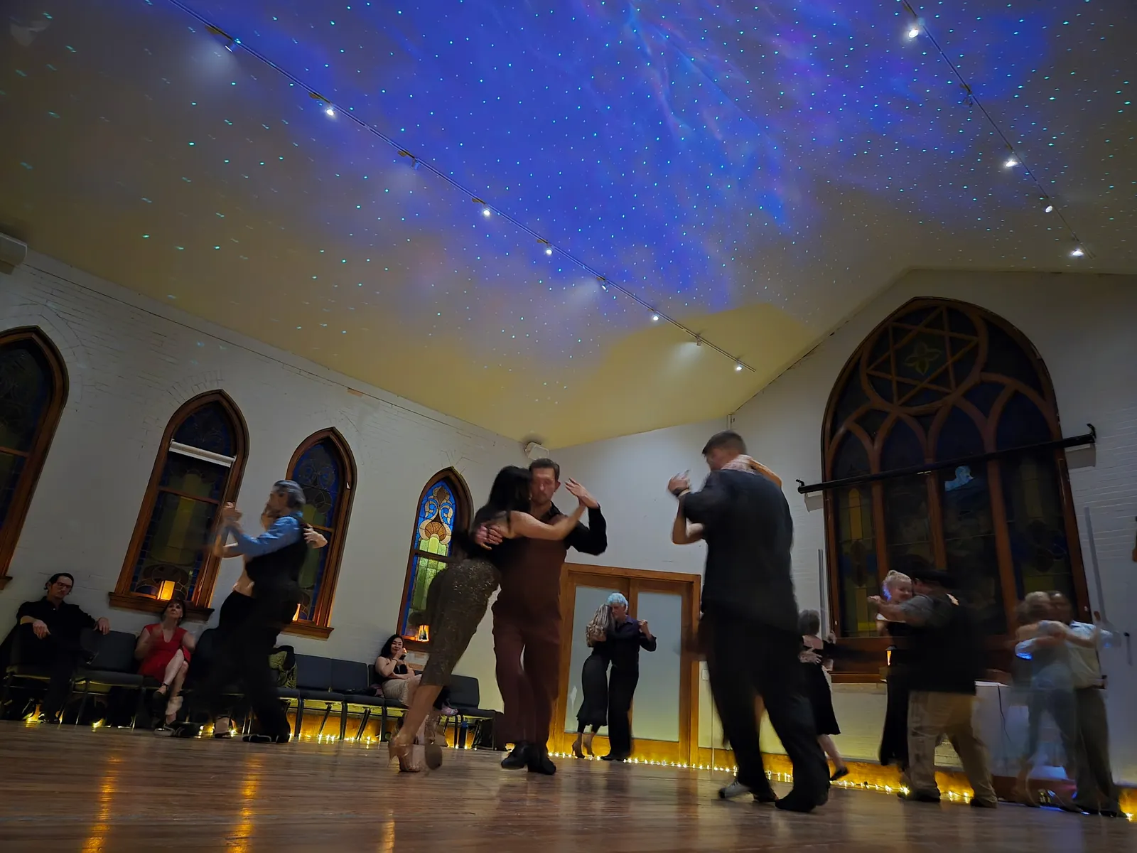 Couples dance tango beneath a starry projected ceiling, the hall's tall arched window glowing behind them