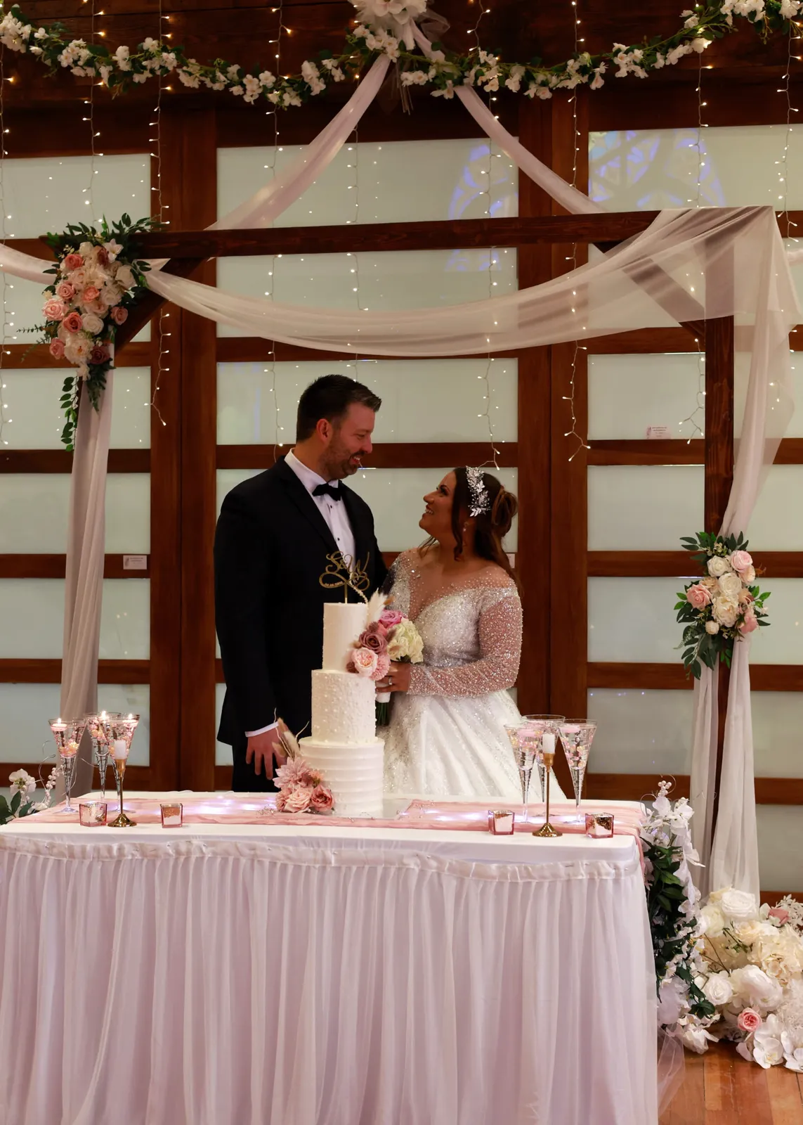 A bride and groom share a look over their wedding cake beneath a draped floral arch in the reception hall