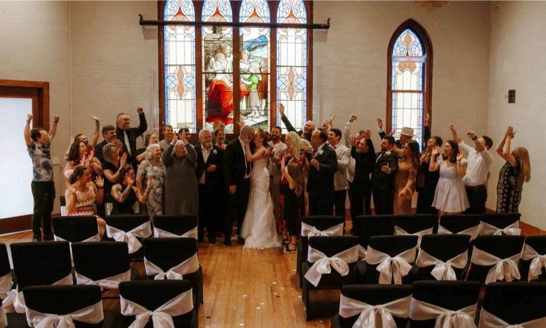 A wedding party cheers as the couple kisses before the grand stained-glass window of the sanctuary