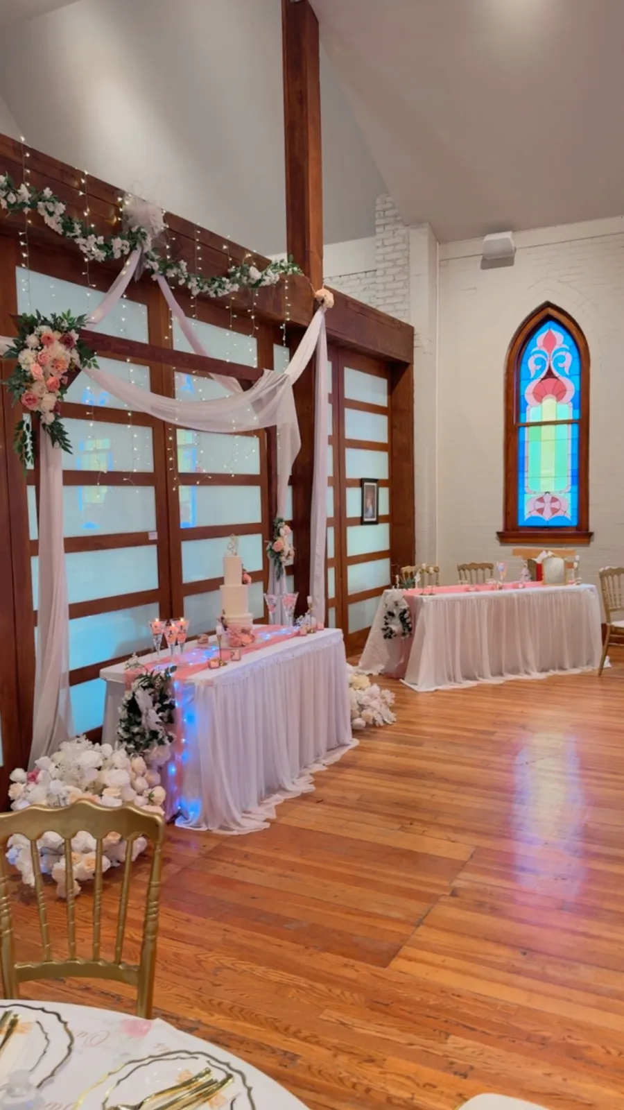 A decorated head table with a tiered wedding cake set against the hall's wooden partition and stained glass