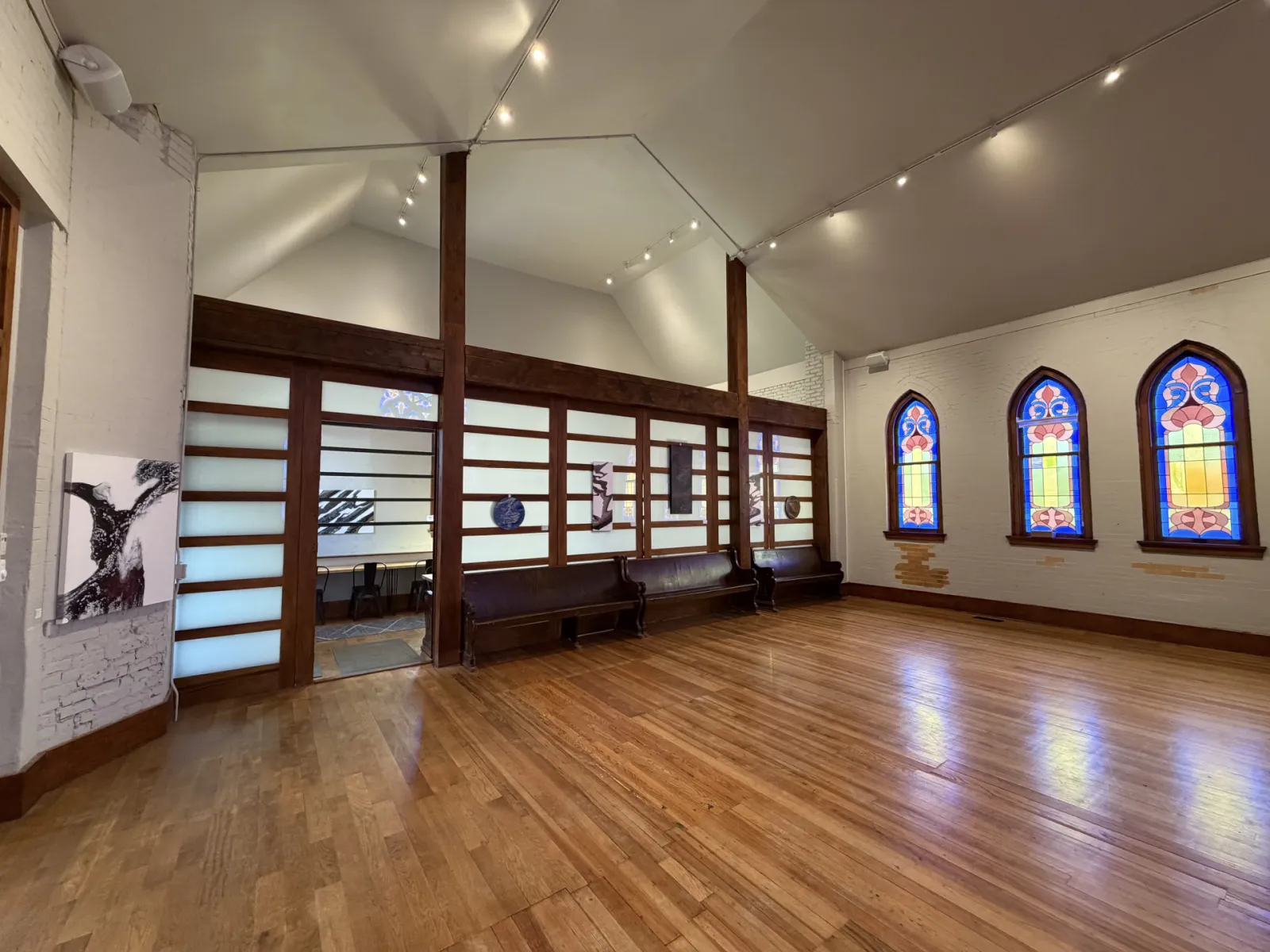 Wide view of the vaulted hall during an art exhibition, with paintings on the white brick and partition walls and stained-glass windows at right