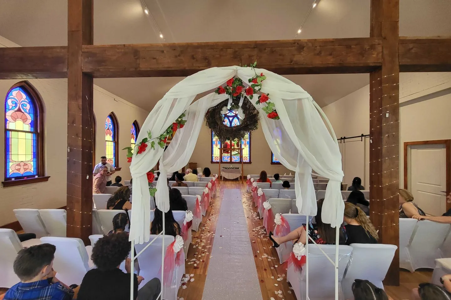 A petal-lined aisle runs beneath a white-draped wedding arch toward the altar as guests take their seats